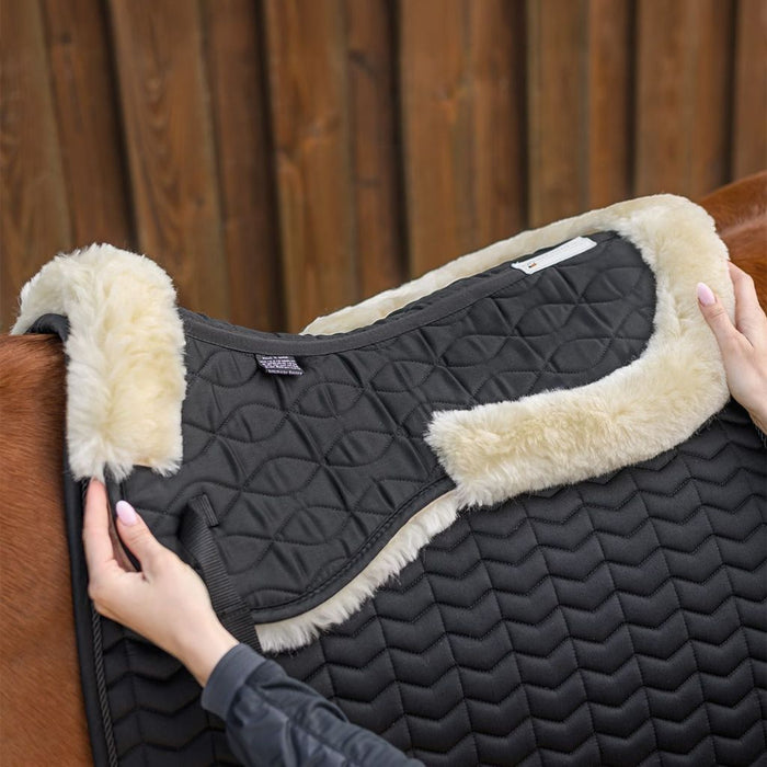 Black quilted saddle pad with beige fleece lining on a horse, against a wooden fence background.